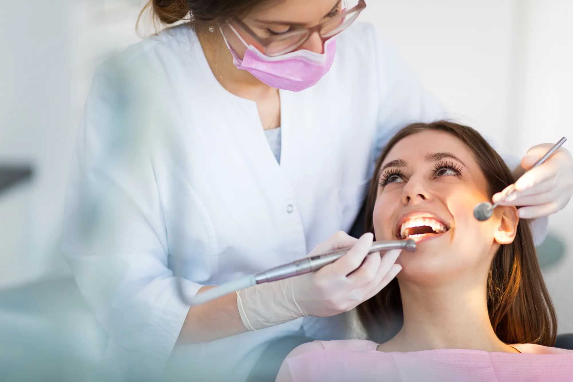 Smiling woman in a dental chair while a dentist in a pink mask examines her teeth